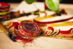 قوطی فلزی گرد صدف پک با طرح سنتی بته جقه قرمز، حاوی زعفران صادراتی، در کنار کاغذ خوشنویسی و قلم نی